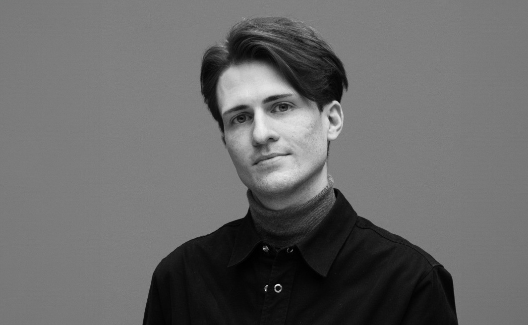 En person med kort mörkt hår och klädd i en svart skjorta och polotröja står mot en enkel bakgrund.