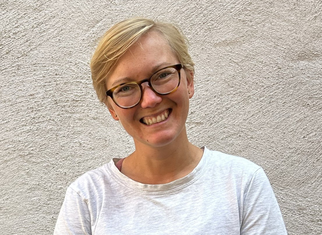 En person med kort blont hår och glasögon ler mot kameran, klädd i en ljus skjorta. Bakgrunden är en strukturerad ljus vägg.