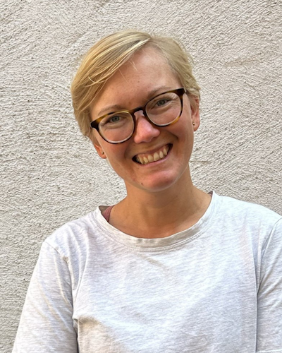 En person med kort blont hår och glasögon står mot en ljus strukturerad vägg. Personen ler mot kameran och har på sig en ljusgrå skjorta.