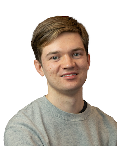 En person med kort brunt hår och en grå tröja ler mot kameran framför en vit bakgrund.