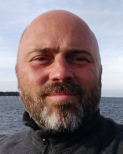En person med skägg i en grå jacka tar en selfie vid havet, med horisonten och molnig himmel i bakgrunden.