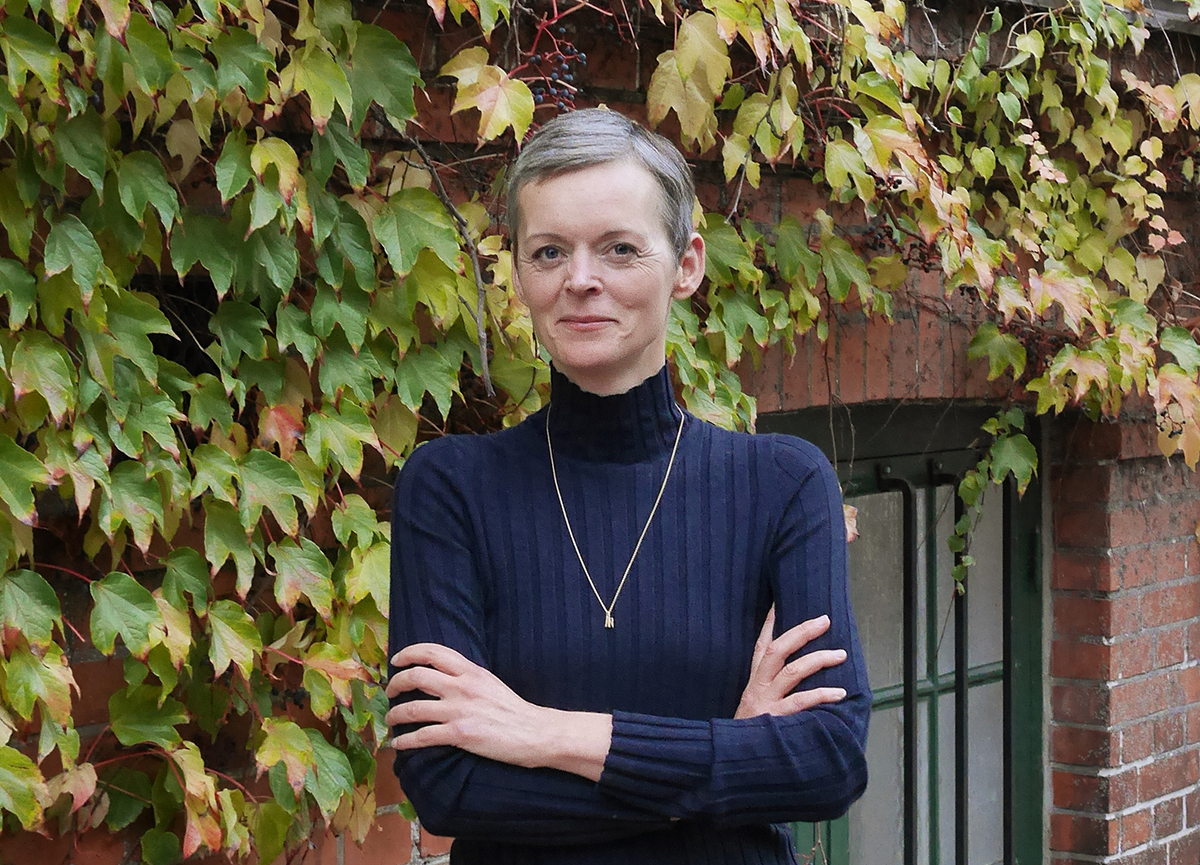 En person med kort hår, klädd i en mörk polotröja och ett halsband, står med armarna i kors framför en vägg täckt av gröna och bruna murgrönsblad.