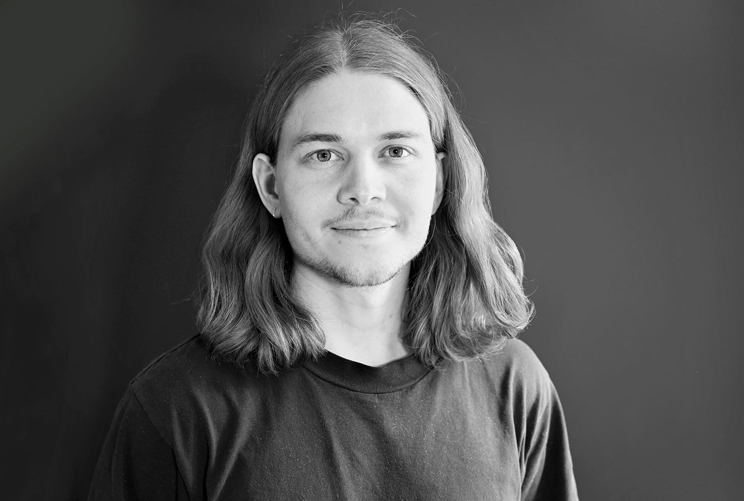 En person med långt hår, iklädd och mörk skjorta, står framför en mörk bakgrund.