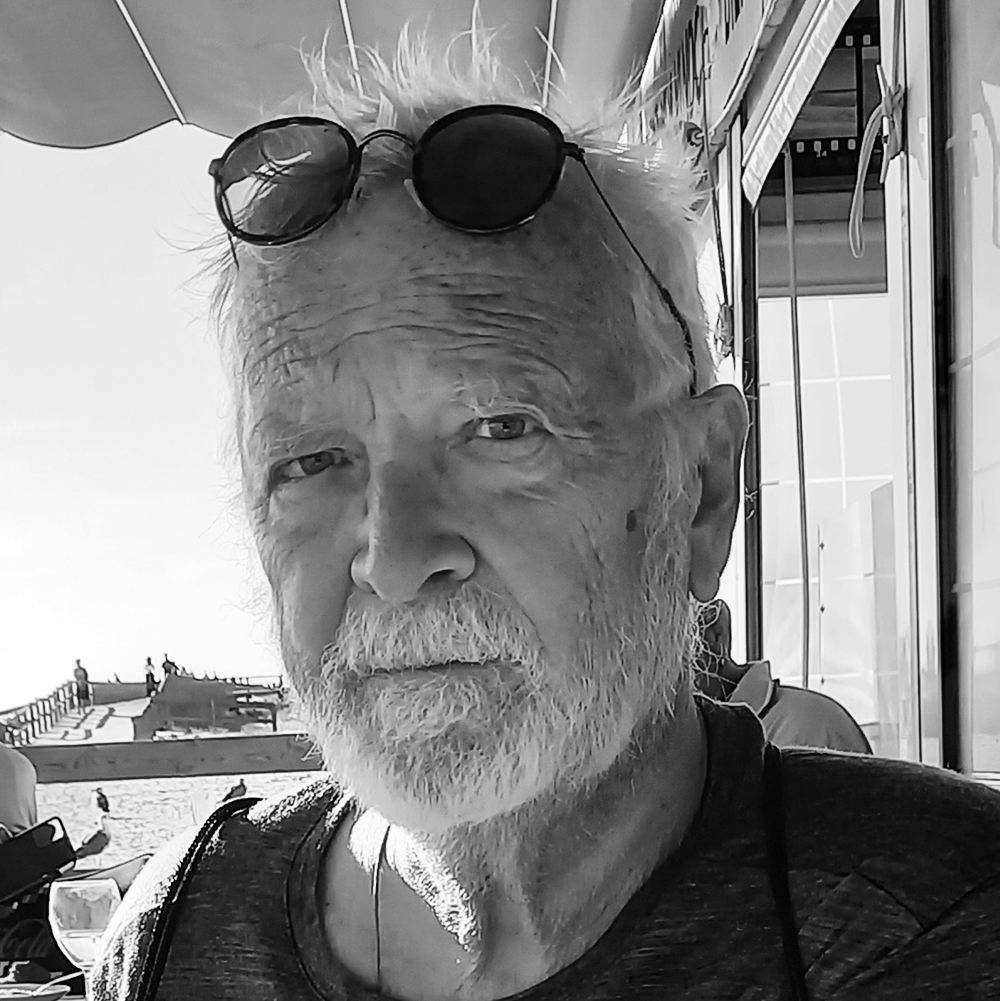 En man med glasögon på huvudet och en T-shirt tittar in i kameran medan han sitter utomhus nära en strand.