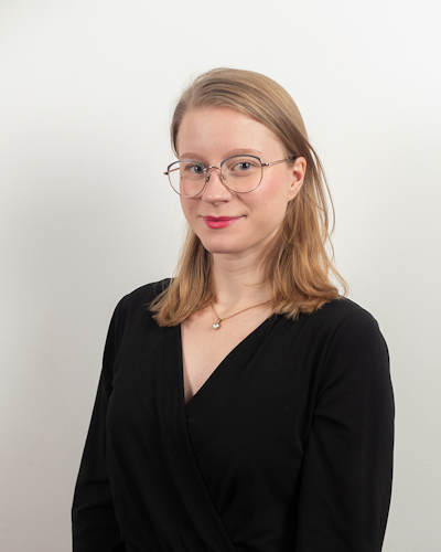 En person med glasögon och axellångt hår har på sig en svart tröja och står mot en vit bakgrund.