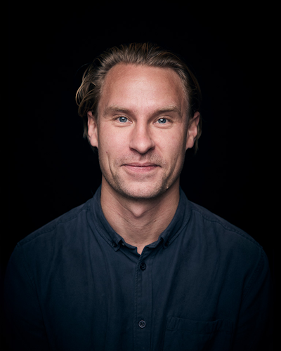 En person med blont hår uppsatt, iklädd och skjorta med knappar, tittar rakt mot kameran mot en mörk bakgrund.