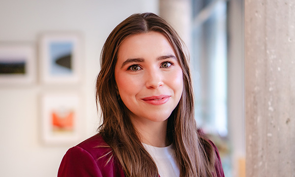 En person med brunt långt hår och en mörkröd jacka står inomhus och ler försiktigt. I bakgrunden syns suddiga inramade bilder.