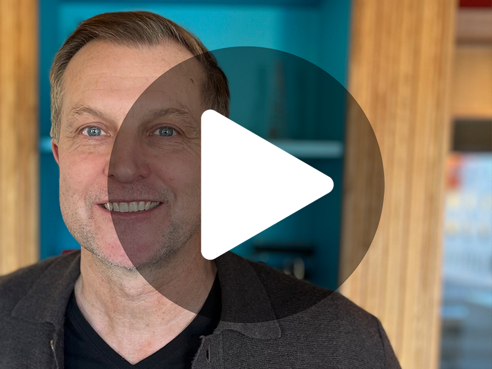 En leende man med kort hår i en mörk skjorta står inomhus; en genomskinlig ikon av en spelknapp visa över bilden.