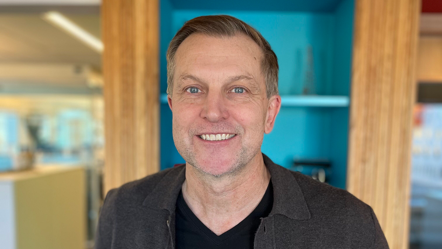 En man ler inomhus med hyllor och en ljusblå vägg i bakgrunden.