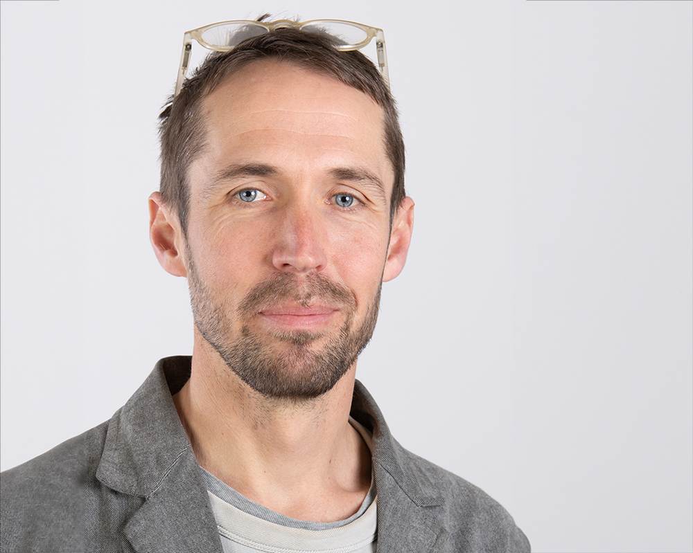 En man med kort hår och skägg, bär glasögon på huvudet, en grå blazer och en grårandig skjorta, står mot en vit bakgrund.