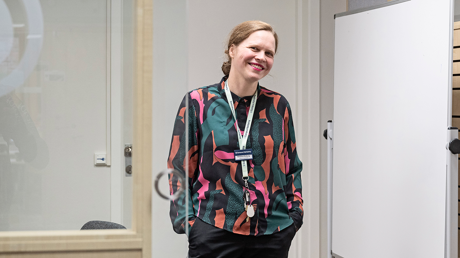 En person står i ett kontor, klädd i skjorta och har en namnbricka runt halsen, ler mot kameran.