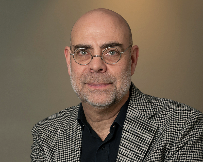 En flintskallig man med glasögon och skägg har på sig en rutig kavaj över en svart skjorta. Han är vänd mot kameran framför en enkel bakgrund.