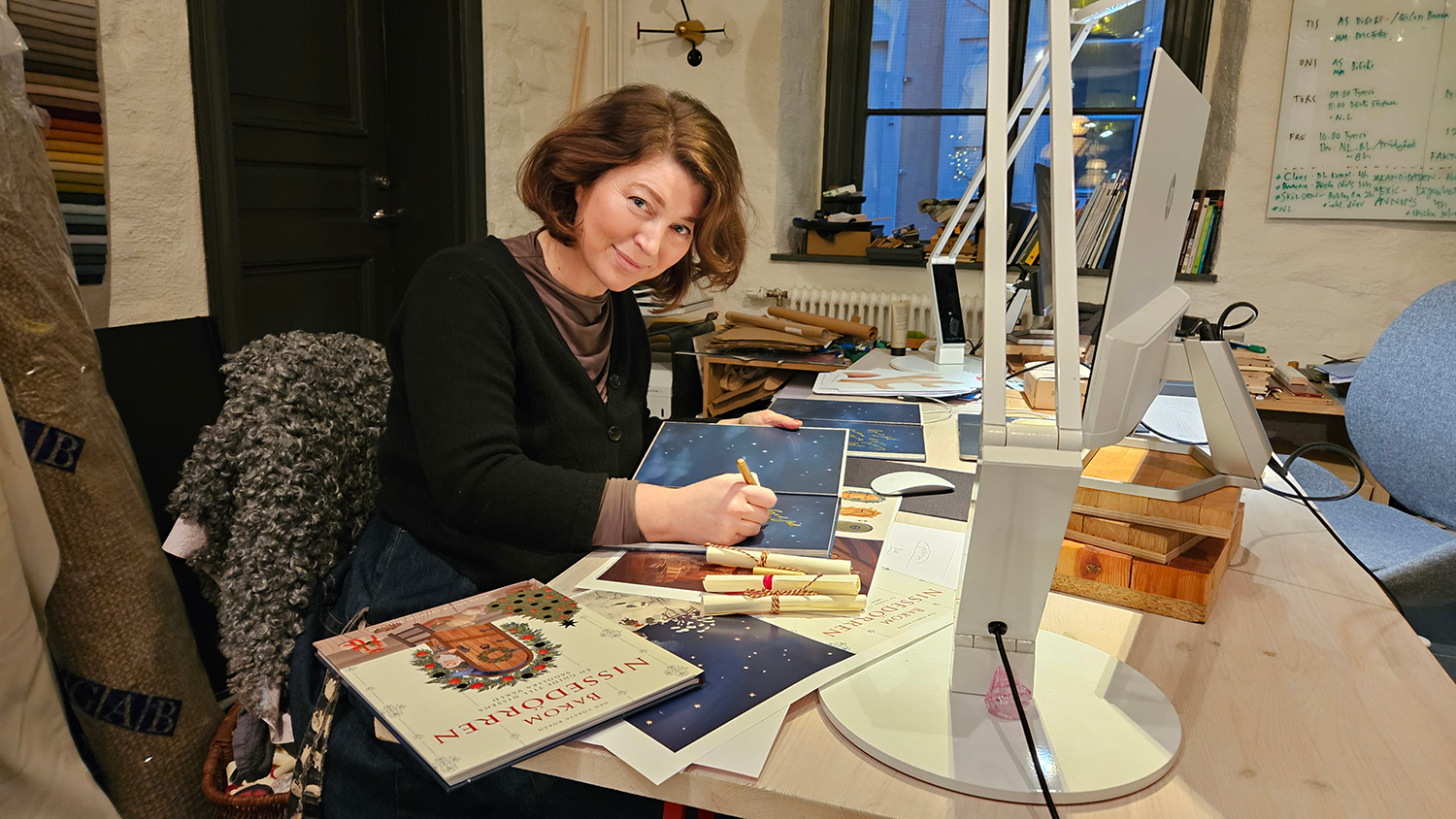 En person sitter vid ett skrivbord på kontoret med böcker och papper runt sig och ler medan han håller en penna i handen nära flera datorskärmar.