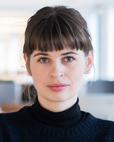 En person med kort brunt hår och lugg, klädd i en svart polotröja, tittar rakt in i kameran. Bakgrunden är ett suddigt inomhusmiljö.