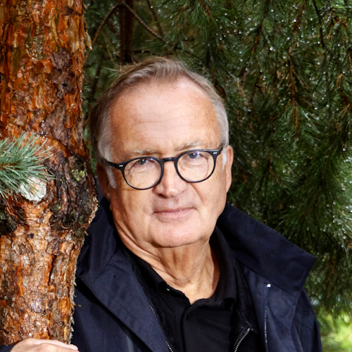 En äldre man med glasögon och en mörk jacka står bredvid ett träd i ett skogsområde.