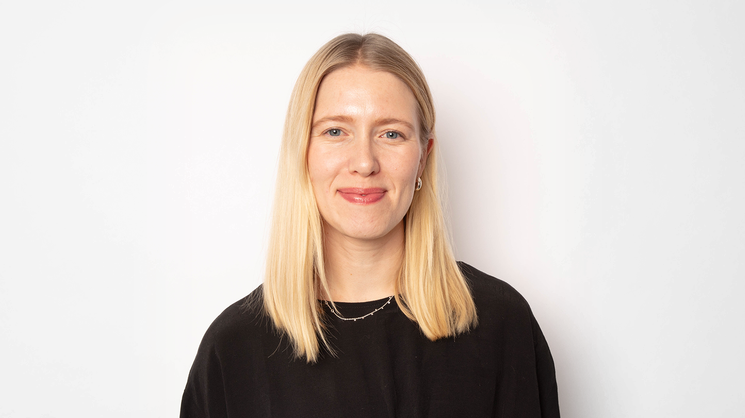 En kvinna med blont hår, klädd i svart, står mot en vit bakgrund och ler lite.