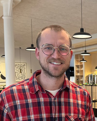 En person med glasögon och en rutig röd skjorta ler i ett upplyst rum med hängande lampor och abstrakt konst på väggen.