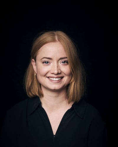 En person med röd axellångt hår ler mot en svart bakgrund och har på sig en mörk tröja.