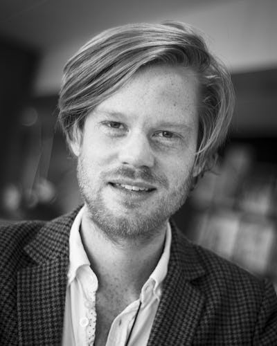 Svartvit bild av en person med medellångt hår och skägg, klädd i en rutig kavaj och vit skjorta, som tittar mot kameran.