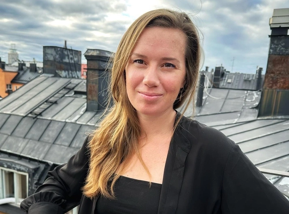 Kvinna med långt hår står på ett tak med molnig himmel och stadens byggnader i bakgrunden.