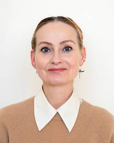 En person med beige tröja och en vit skjorta och står mot en enkel bakgrund.