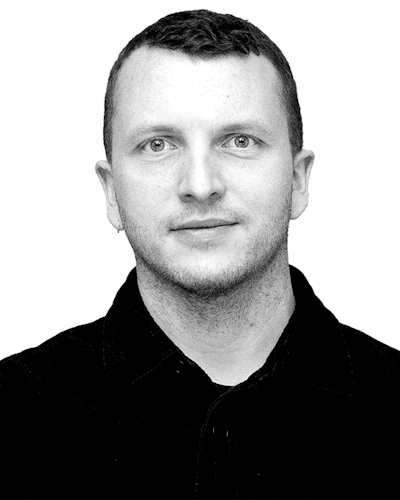 Svartvitt porträtt av en person med kort hår, iklädd en mörk skjorta och vänd mot kameran framför en enkel bakgrund.