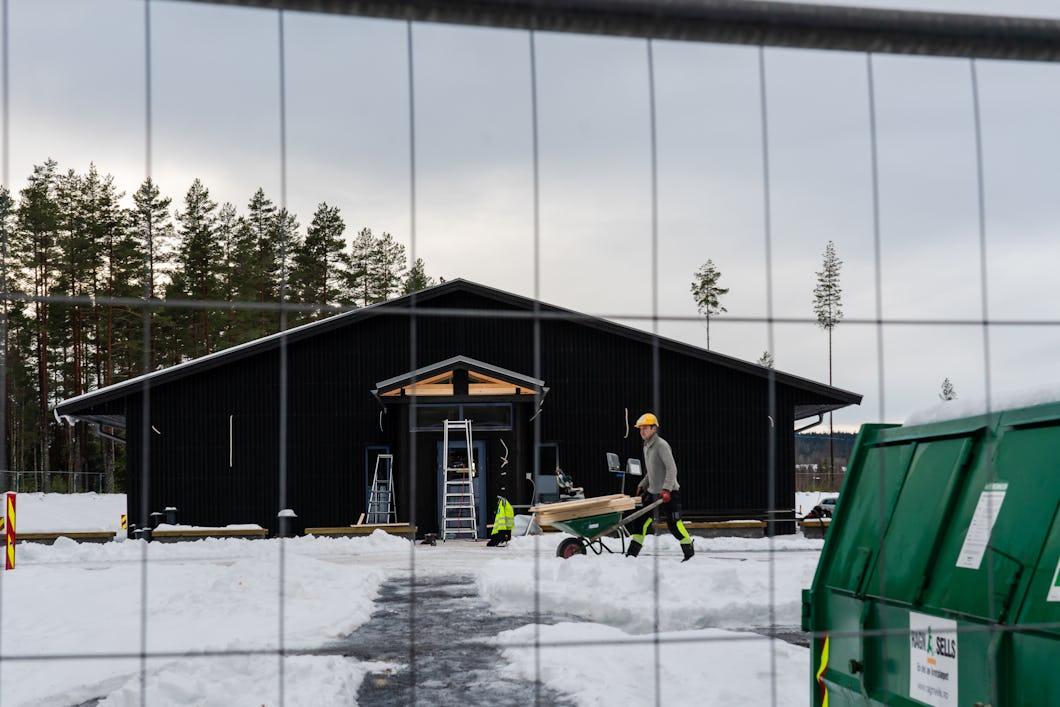 En byggarbetare skjuter en skottkärra framför en mörk byggnad omgiven av snö, satt genom ett staket. Stegar och fler arbetare syns vid ingången till byggnaden.