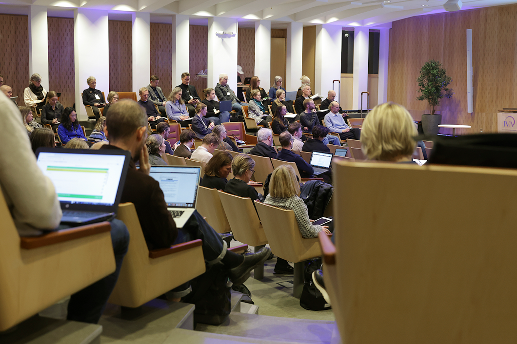 Personer sitter i en lärosal med nivåindelade platser, några använder datorer, medan en talare presenterar längst fram.