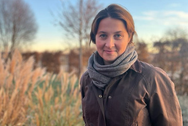 En person i jacka och halsduk står utomhus med högt gräs och träd utan löv i bakgrunden under en klar himmel.