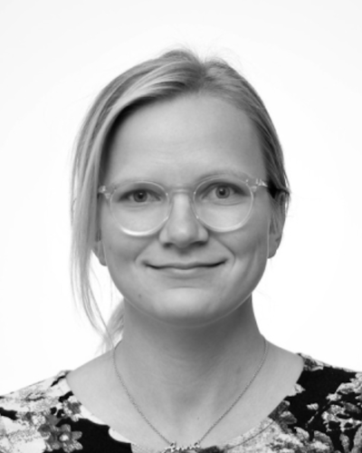 En person som bär glasögon och har axellångt hår, iförd en mönstrad överdel, ler på ett svartvitt porträtt.