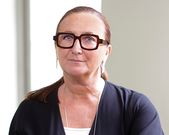 Person med glasögon, i en mörk topp och långa örhängen, står framför en ljus bakgrund.