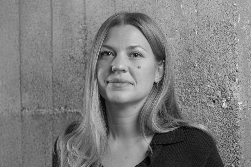 Svartvitt porträtt av en kvinna med långt hår och ett litet leende, ståendes mot en betongvägg med textur.
