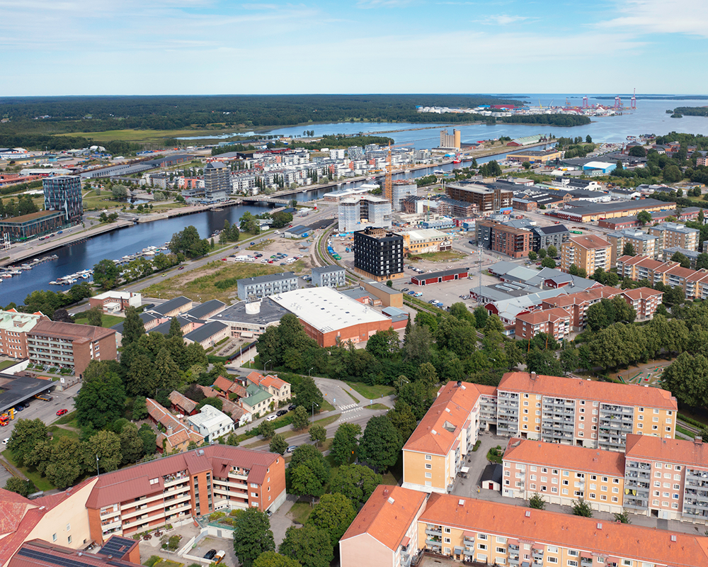 Flygvy över en stad vid kusten med bostadsbyggnader, vattenvägar och grönområden under en molnfri himmel.