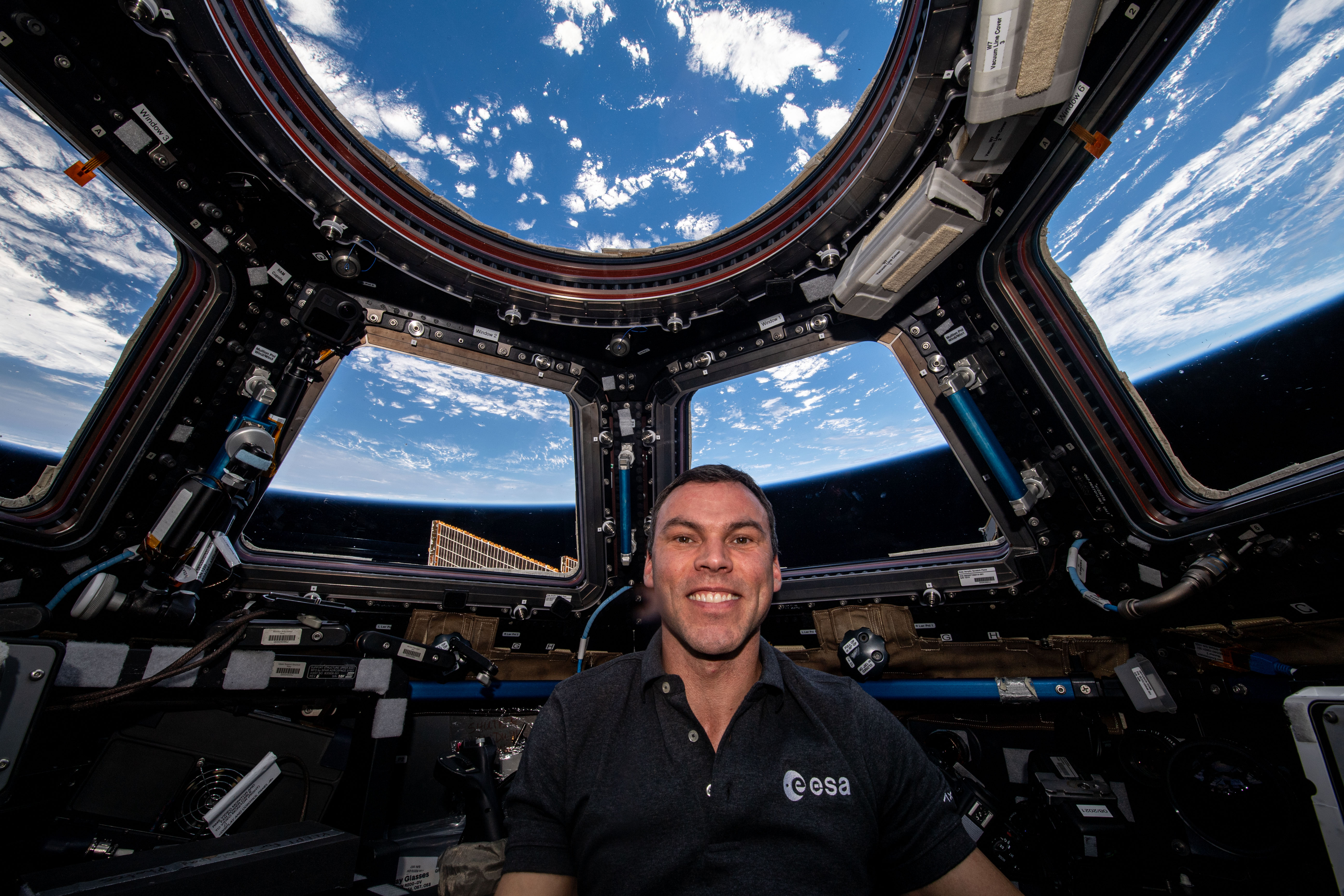 En astronaut inne i kupolen på den internationella rymdstationen ler medan jorden syns genom flera fönster.
