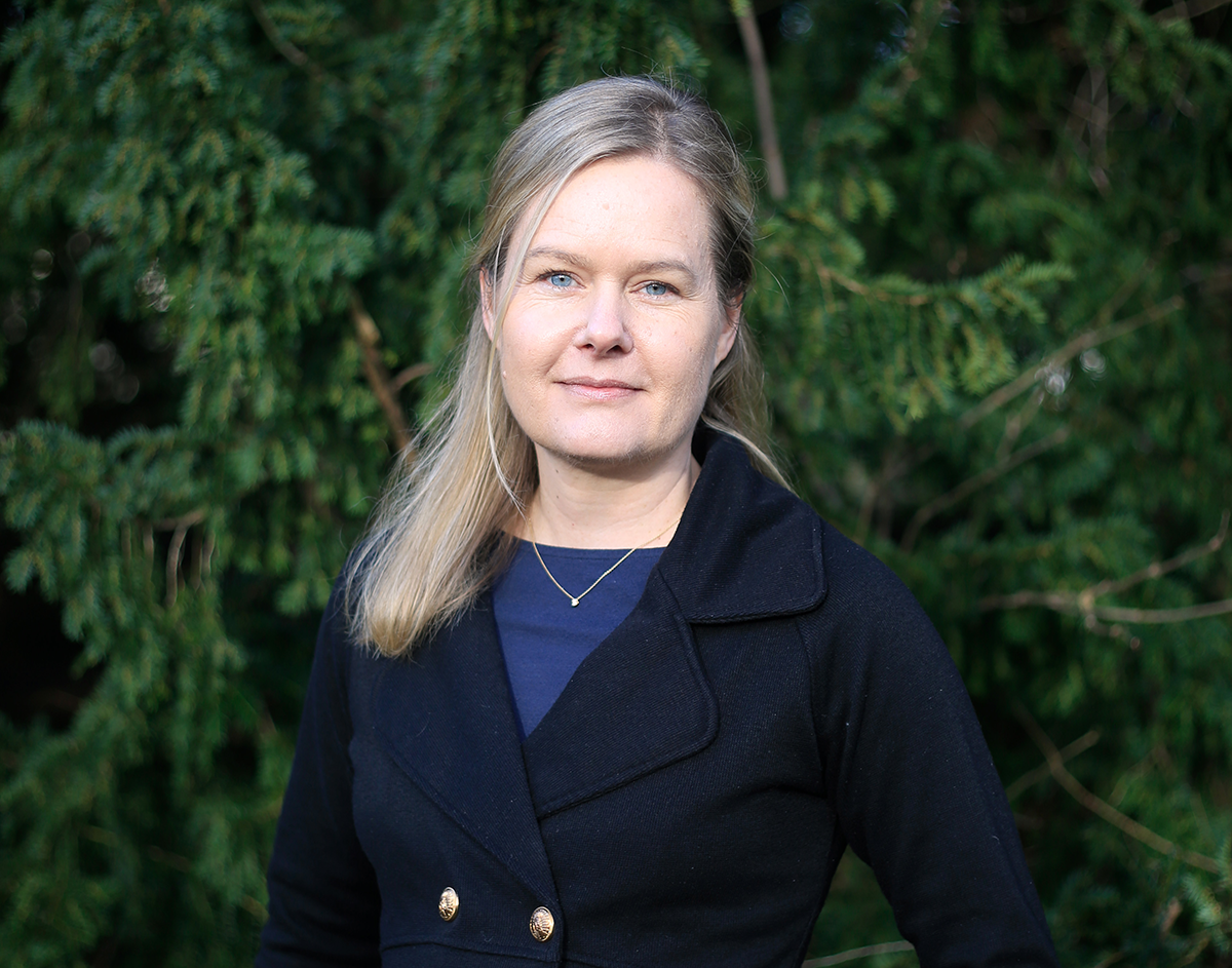 Kvinna med blont hår i en svart rock står framför en bakgrund av växter.