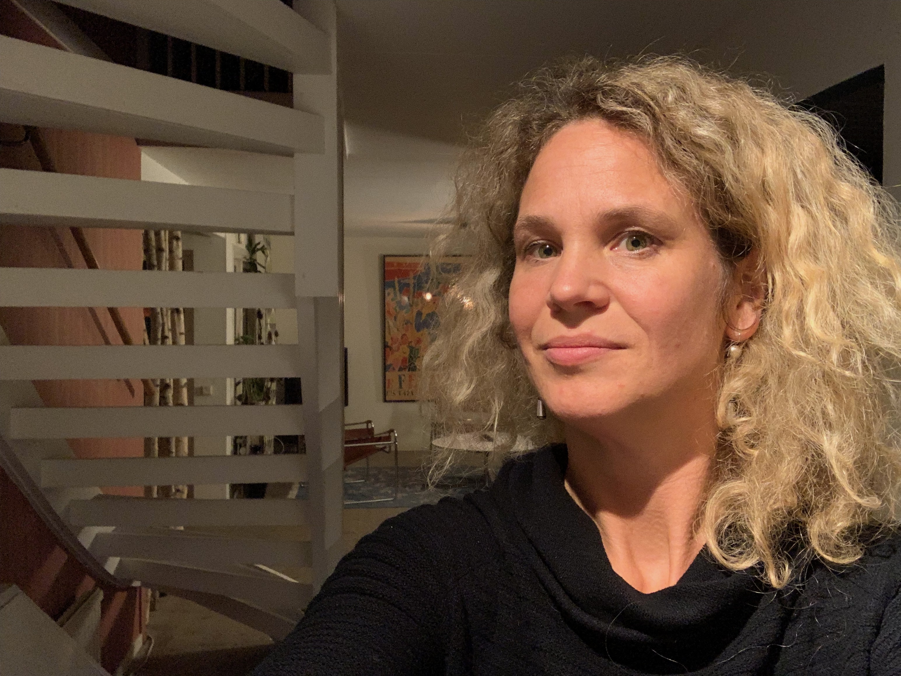 En kvinna med lockigt blont hår och svart topp tar en selfie inomhus nära en vit trappa.
