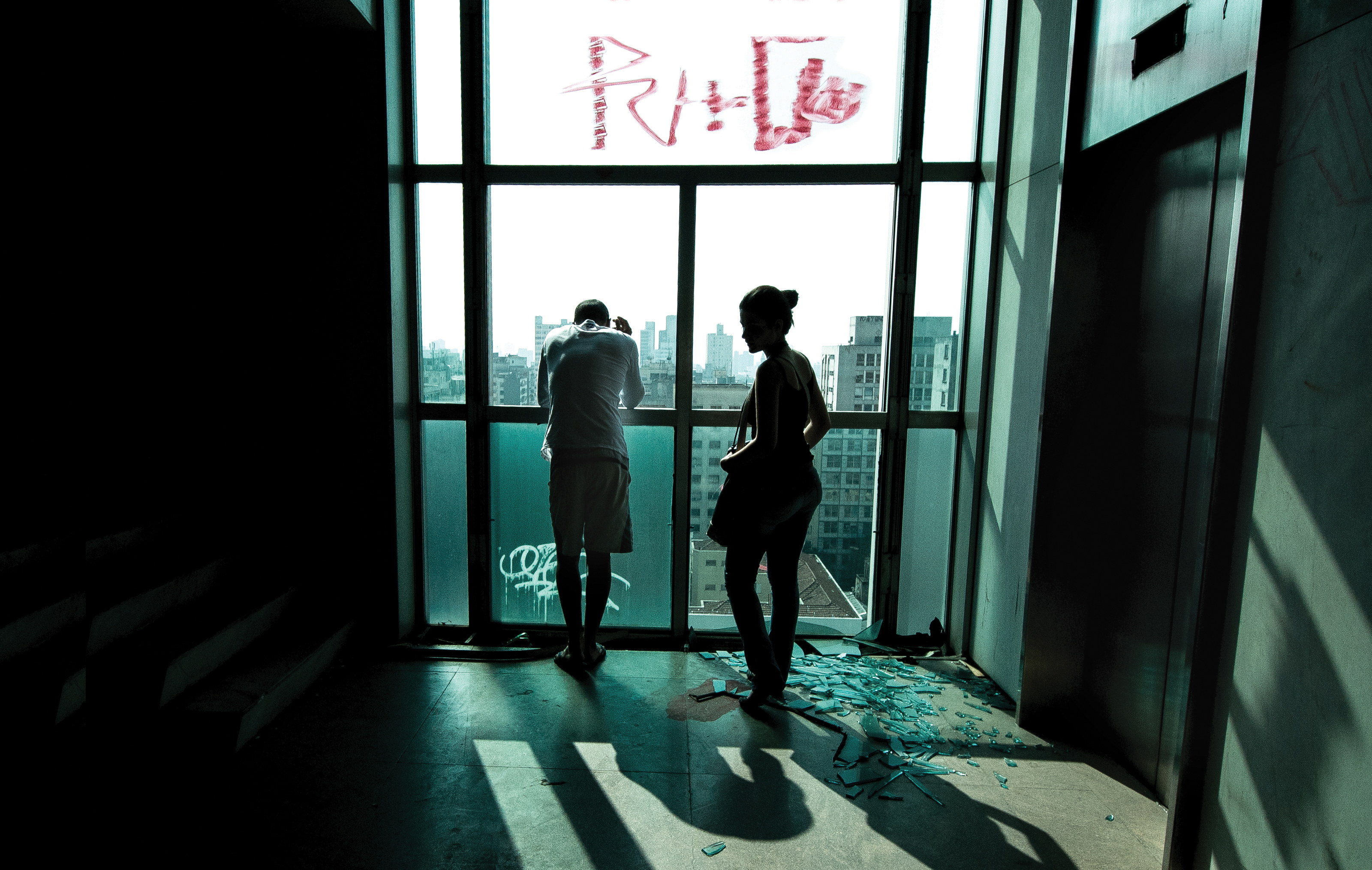 Två personer står vid stora fönster i ett rum med trasigt glas på golvet och klotter på fönstret, med utsikt över en stad.