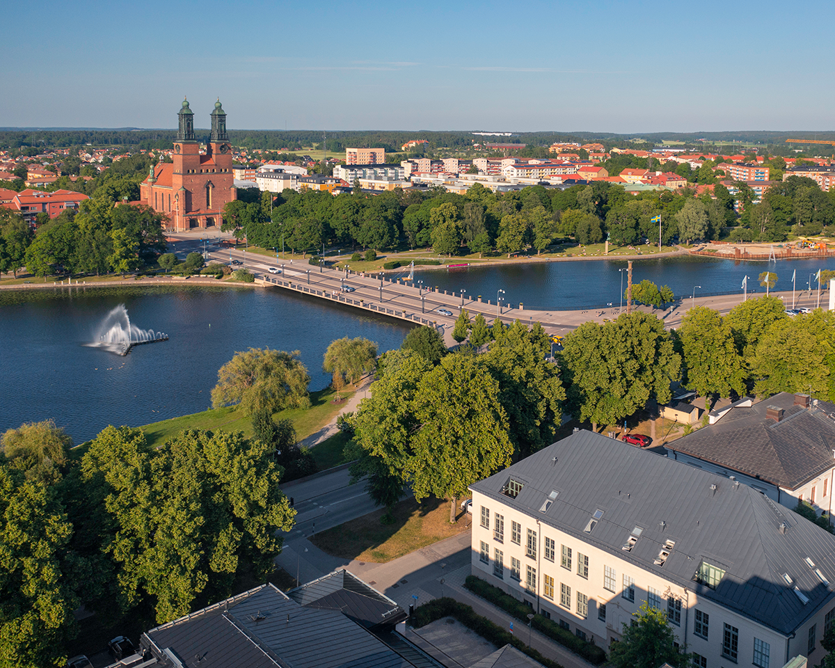 Flygbild över en stad med en stor kyrka, en bro över en flod och en fontän. Träd och byggnader omger området under en molnfri himmel.