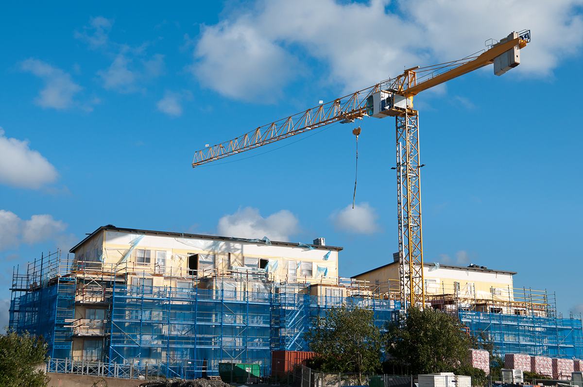 Byggarbetsplats med en stor gul kran och en byggnad täckt av blått byggnadsställning, mot en himmel med moln.