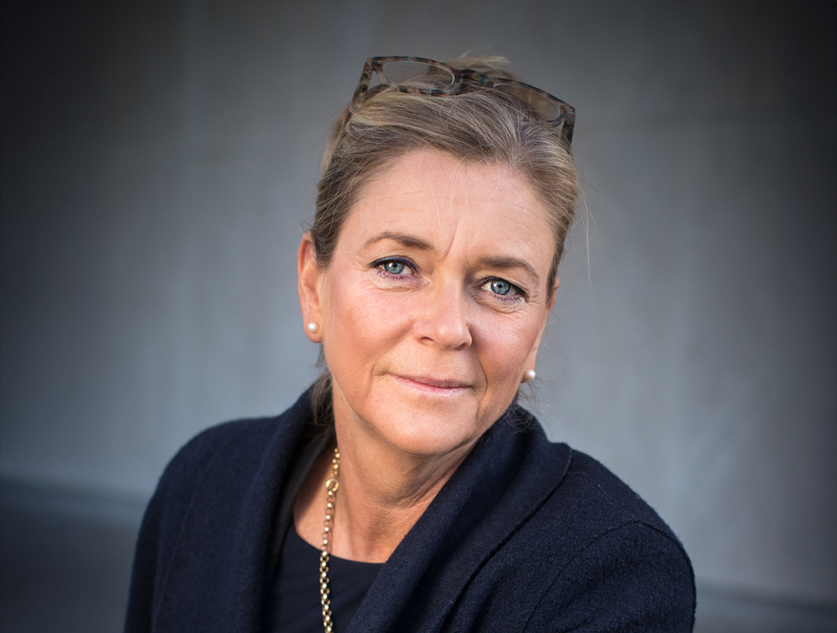 En person med kort hår som har glasögon på huvudet, örhängen, en mörk kavaj och ett halsband, tittar rakt in i kameran.