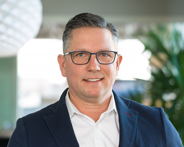 En man med kort mörkt hår och glasögon, iklädd en vit skjorta och en mörk blazer, står inomhus med en suddig bakgrund.