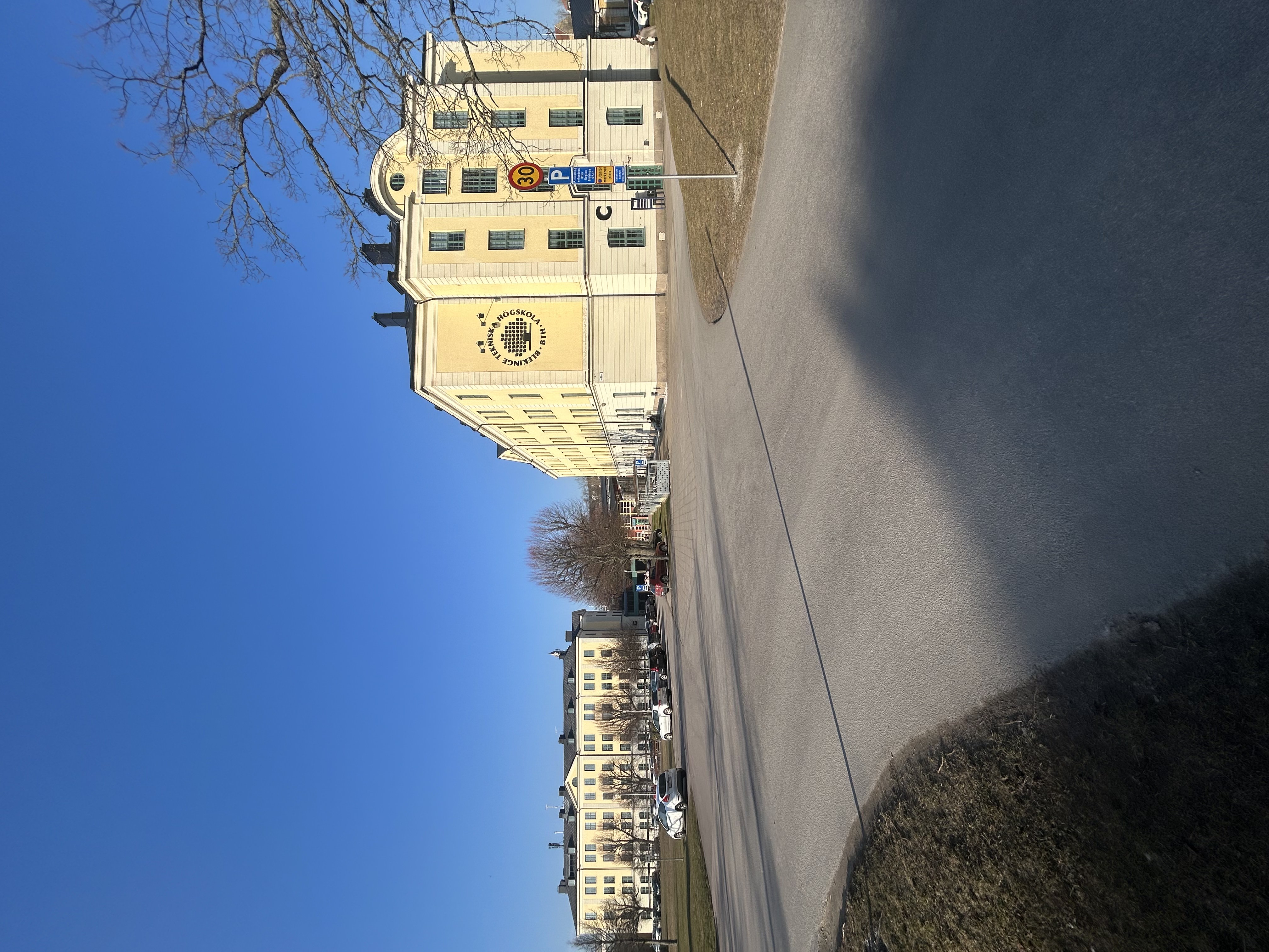 Två flerfamiljshus i gul färg står under en klar himmel, med en väg i förgrunden och träd utan löv i närheten.