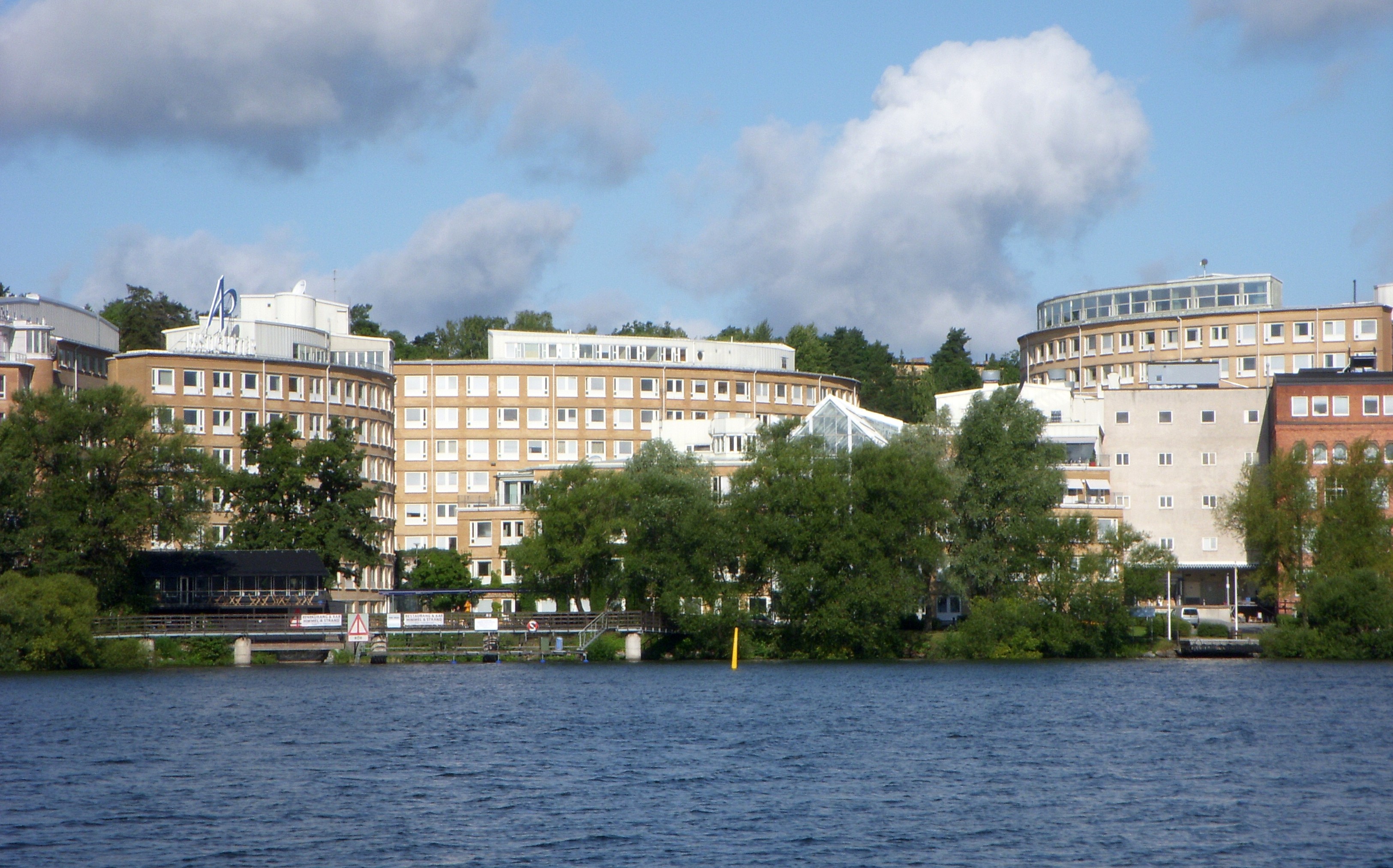 Utsikt mot flera sammanhängande byggnader vid vattnet, omgivna av träd, under en delvis molnig himmel.