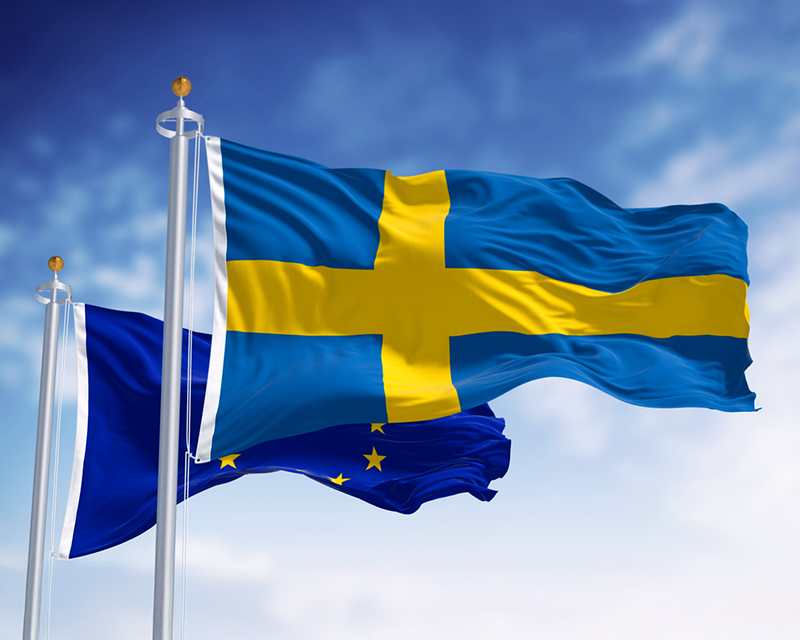 Den svenska flaggan och EU-flaggan vajar på flaggstänger mot en blå himmel.