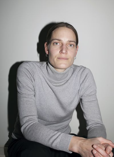 En person sitter mot en enkel vägg, iklädd en ljusgrå polotröja och tittar rakt in i kameran.