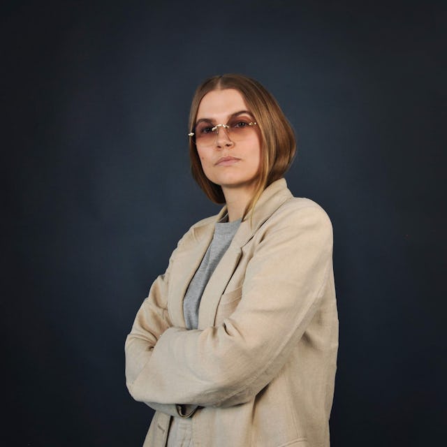 En person med axellångt hår och glasögon, iklädd en beige blazer, står med armarna i kors mot en mörk bakgrund.