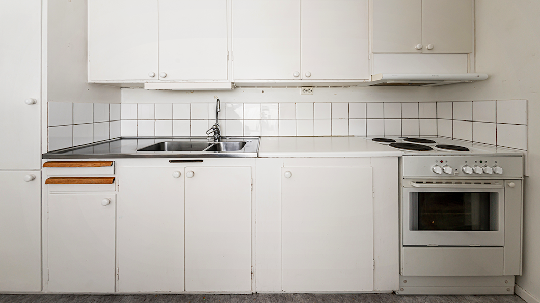 En mindre kök med vita skåp, en diskho i rostfritt stål, en elektrisk spis med fyra plattor och vitt kakel bakom arbetsytan.