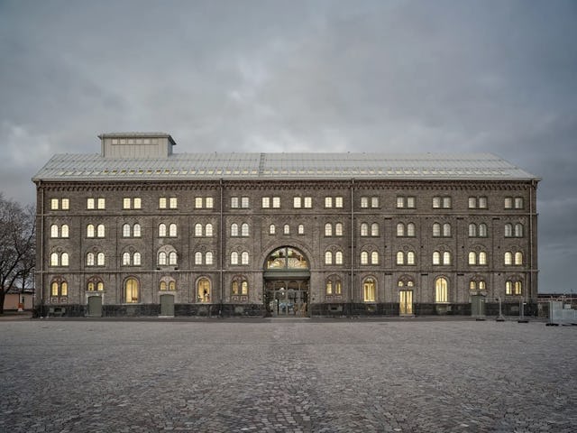 En fyravånings byggnad av sten med rektangulär form och upplysta fönster. Den har ett centralt valv och omges av en gård med gatstenar under en molnig himmel.
