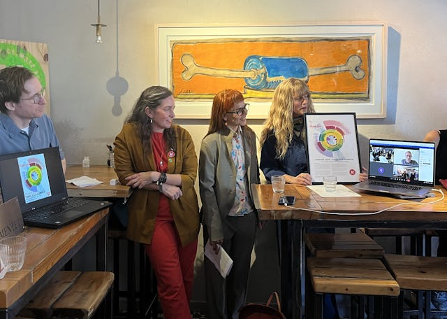Fyra personer står inomhus, två av dem håller en affisch med ett diagram i form av en cirkel. Två bärbara datorer på ett bord visar ett videosamtal. En målning av ett hundben hänger på väggen bakom dem.
