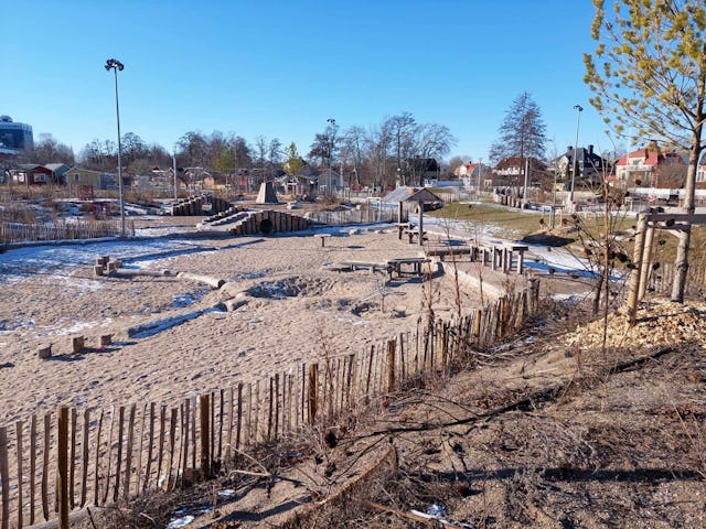 En lekplats utomhus med sand, trälekredskap, en liten bro och några snöfläckar. Platsen är omgiven av ett staket av trä och träd.